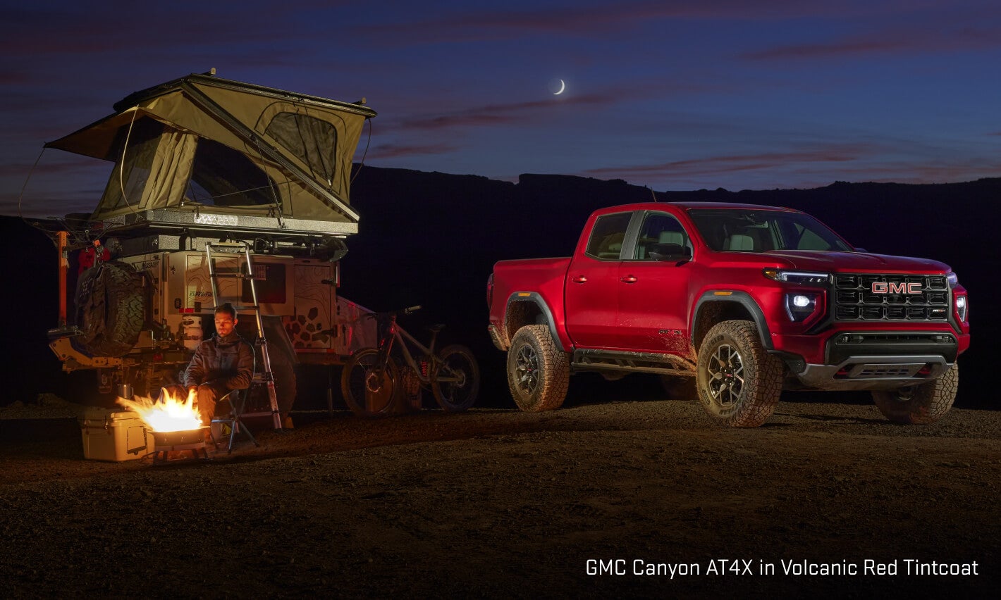 GMC Canyon AT4X exterior in Volcanic Red Tintcoat showing a front three-quarters view parked at a campsite.
